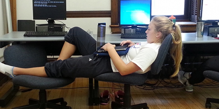 Student sprawled across two chairs, a laptop on her lap and her ponytail hanging over one chair