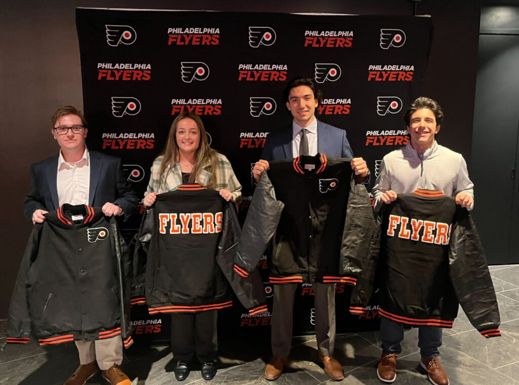 four students pose for a photo at a Flyers game holding jackets they won.